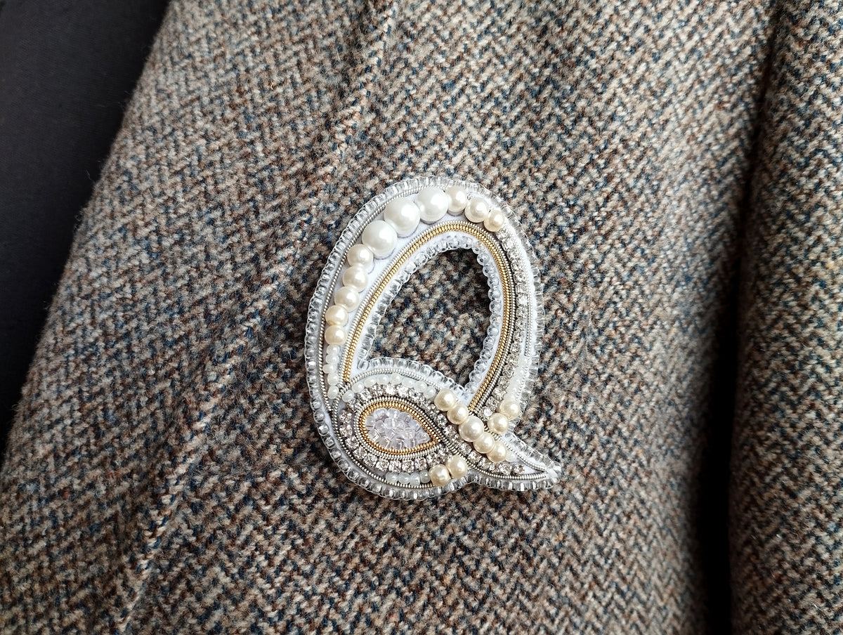 A close-up image of a decorative letter 'Q' pin or brooch on a tweed jacket.