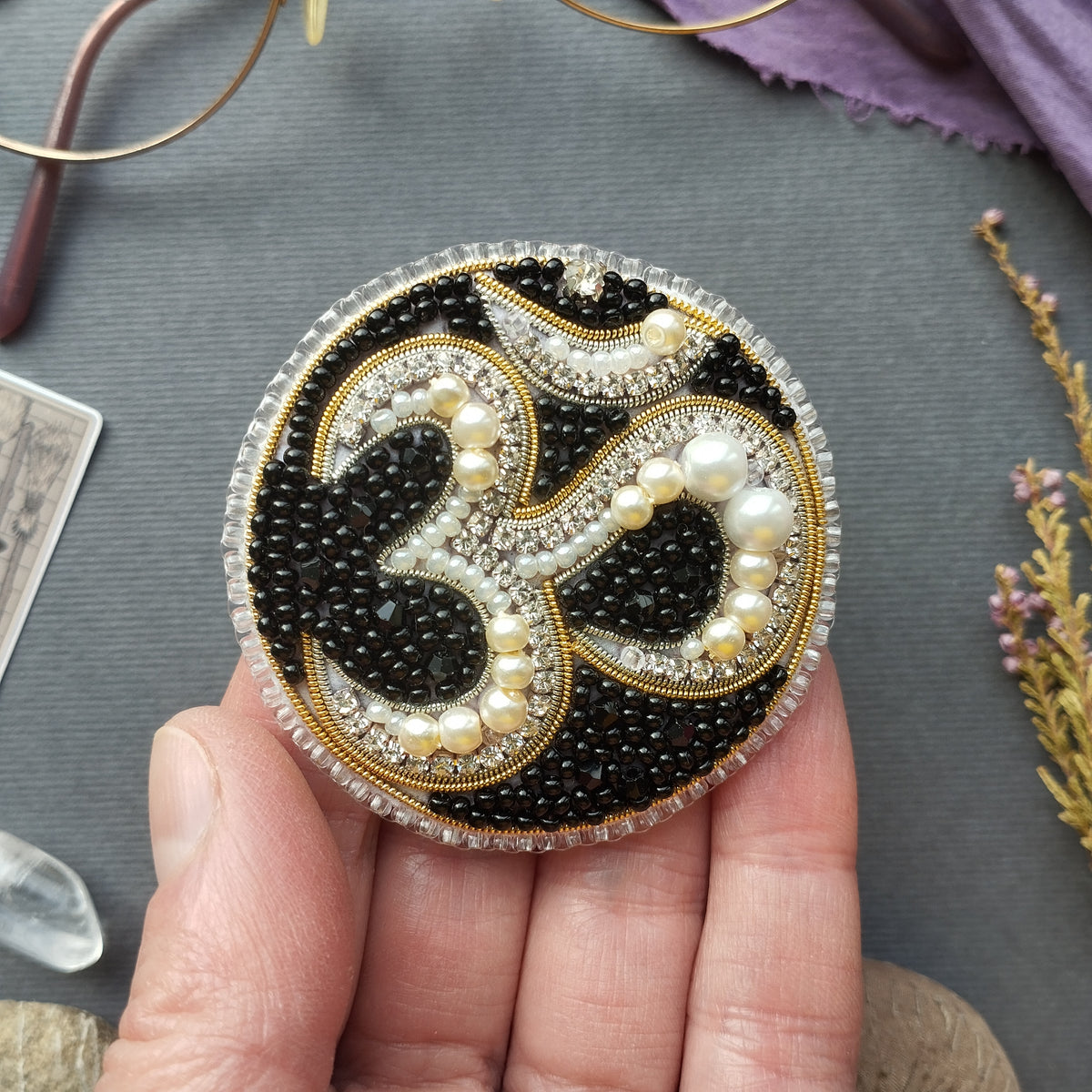A hand holding a circular, ornate piece of jewelry with a black and white design, accented by pearls and rhinestones.