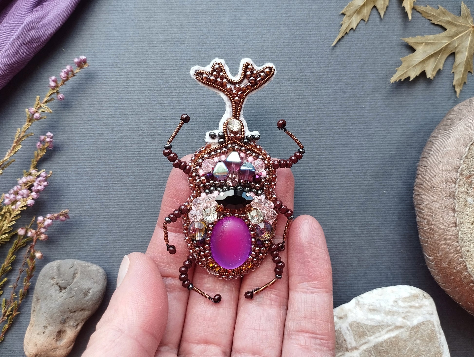 A hand holding a decorative insect brooch with a purple gemstone in the center, surrounded by various natural elements such as leaves, flowers, and rocks.