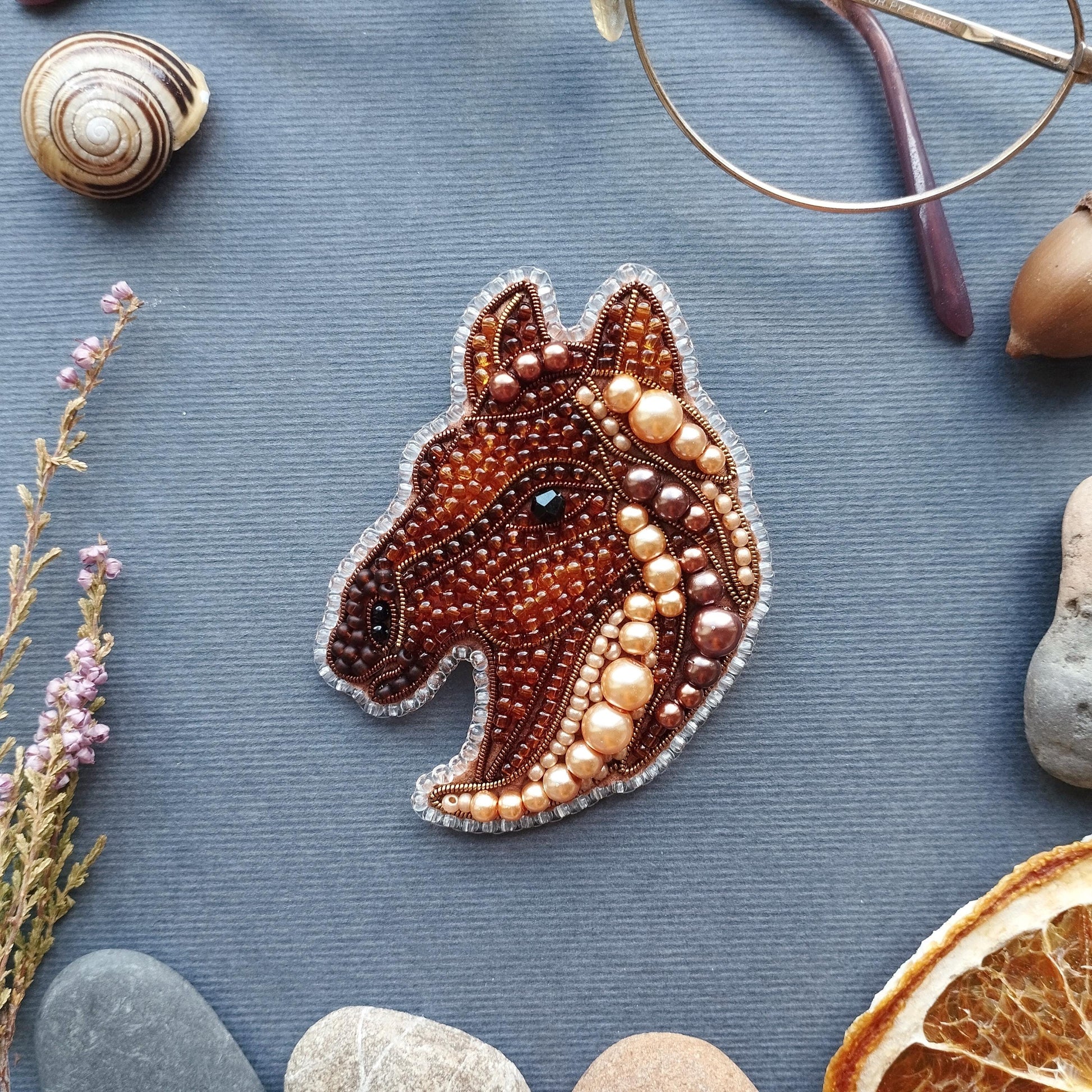 a close-up view of a decorative piece, possibly a brooch or pendant, with a brown and beige color scheme and intricate beadwork. Surrounding the main object are various natural elements, including a seashell, a pair of glasses, a rock, and a slice of orange.