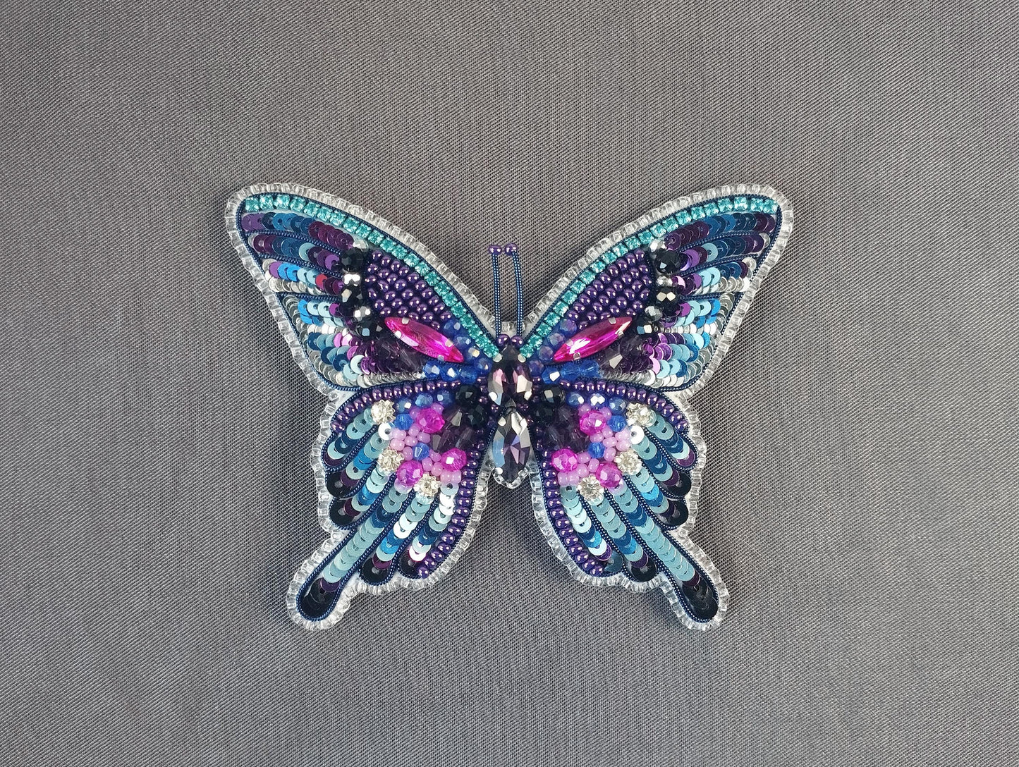A colorful, intricately designed butterfly brooch made of sequins and beads, featuring a vibrant mix of purple, blue, and pink hues.