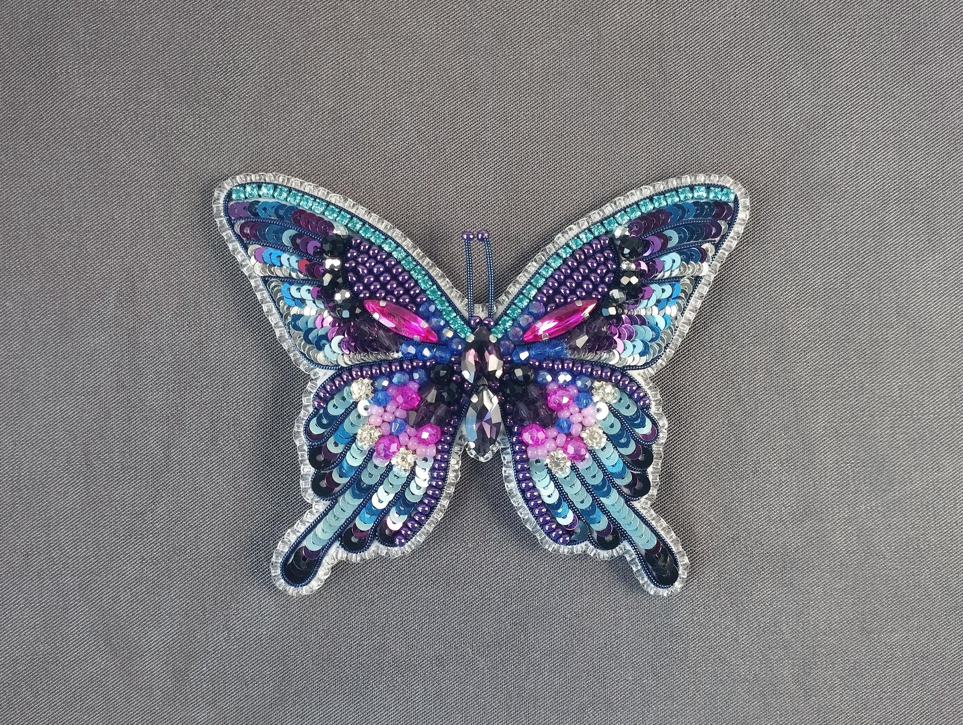 A colorful, intricately designed butterfly brooch made of sequins and beads, featuring a vibrant mix of purple, blue, and pink hues.