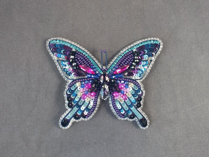 A colorful, intricately designed butterfly brooch made of sequins and beads, featuring a vibrant mix of purple, blue, and pink hues.
