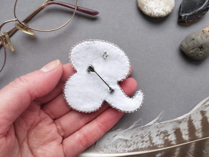 A hand holding a small, white, felt-like object with a silver pin attached to it, surrounded by various objects such as glasses, rocks, and feathers.