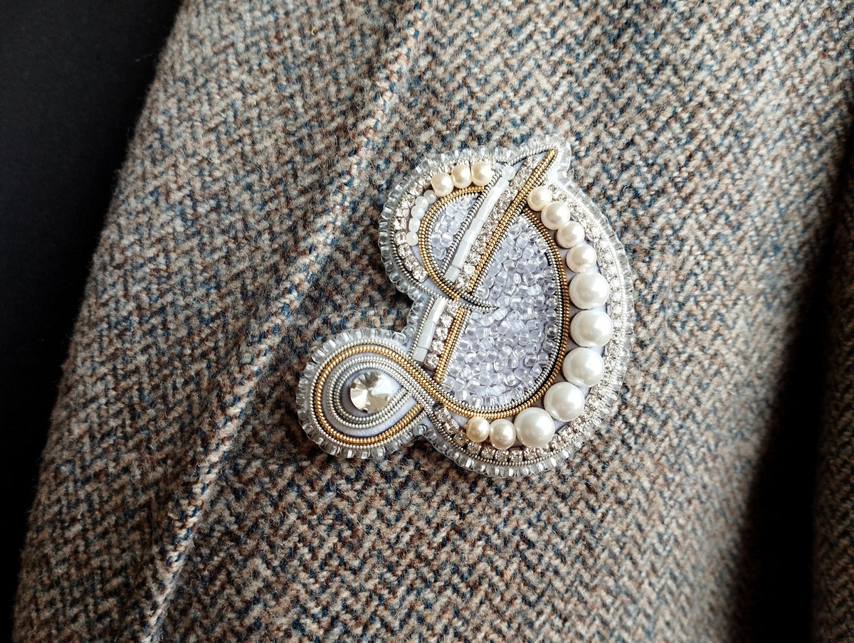 A close-up image of a decorative brooch or pin with a combination of silver, white, and gold elements, including pearls and a floral design.