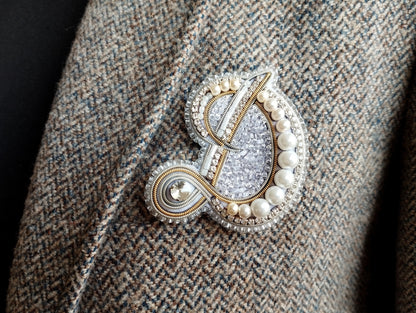 A close-up image of a decorative brooch or pin with a combination of silver, white, and gold elements, including pearls and a floral design.