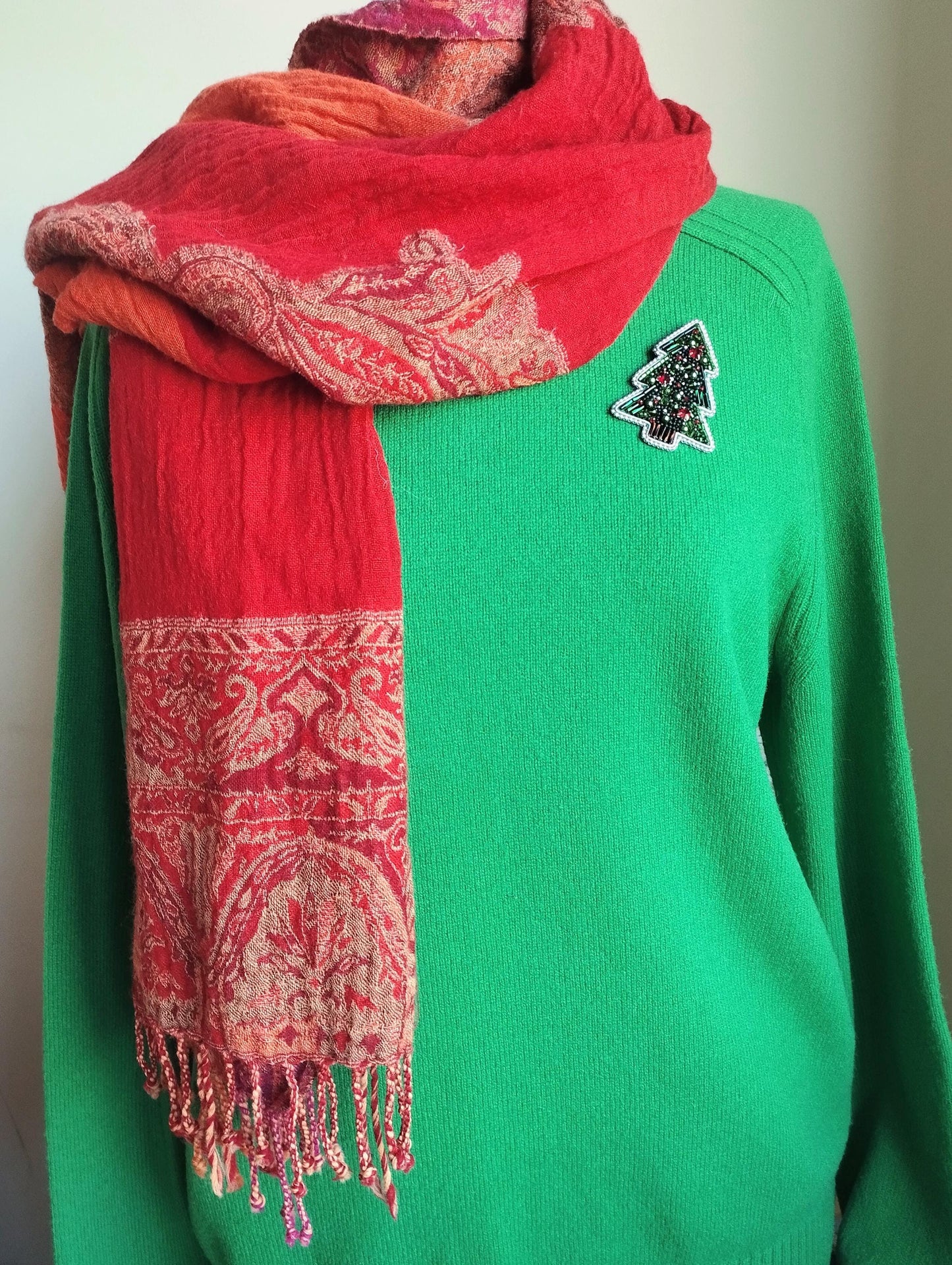 A person is wearing a green sweater with a red and orange patterned scarf draped over it. The scarf has fringe details and a Christmas tree patch on the sweater.