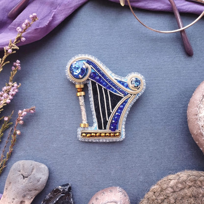a decorative harp brooch with blue and gold accents, placed on a blue surface surrounded by various objects such as a pair of glasses, a plant, and a rock.