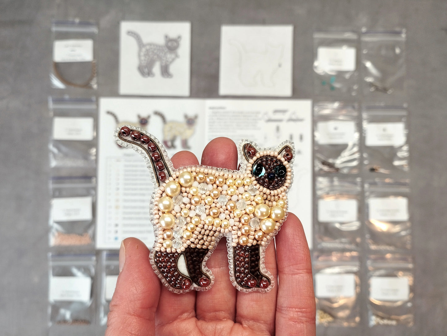 A hand holding a decorative beaded cat pin, with various craft supplies and materials visible in the background.