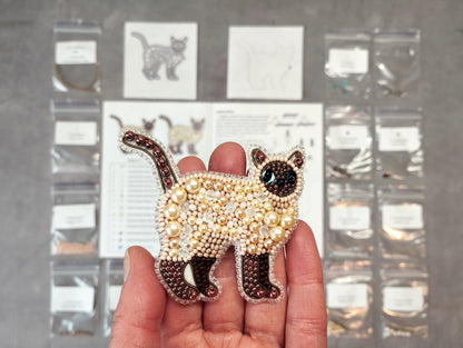 A hand holding a decorative beaded cat pin, with various craft supplies and materials visible in the background.