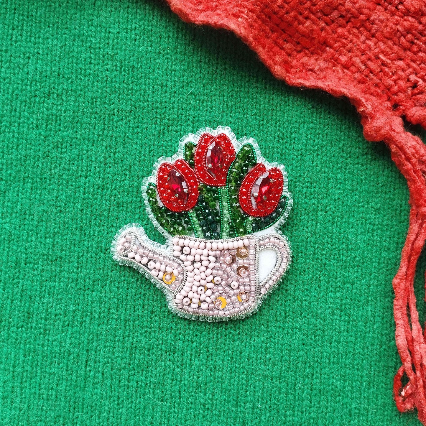 A decorative beaded brooch in the shape of a watering can, adorned with red flowers, is displayed against a green background.