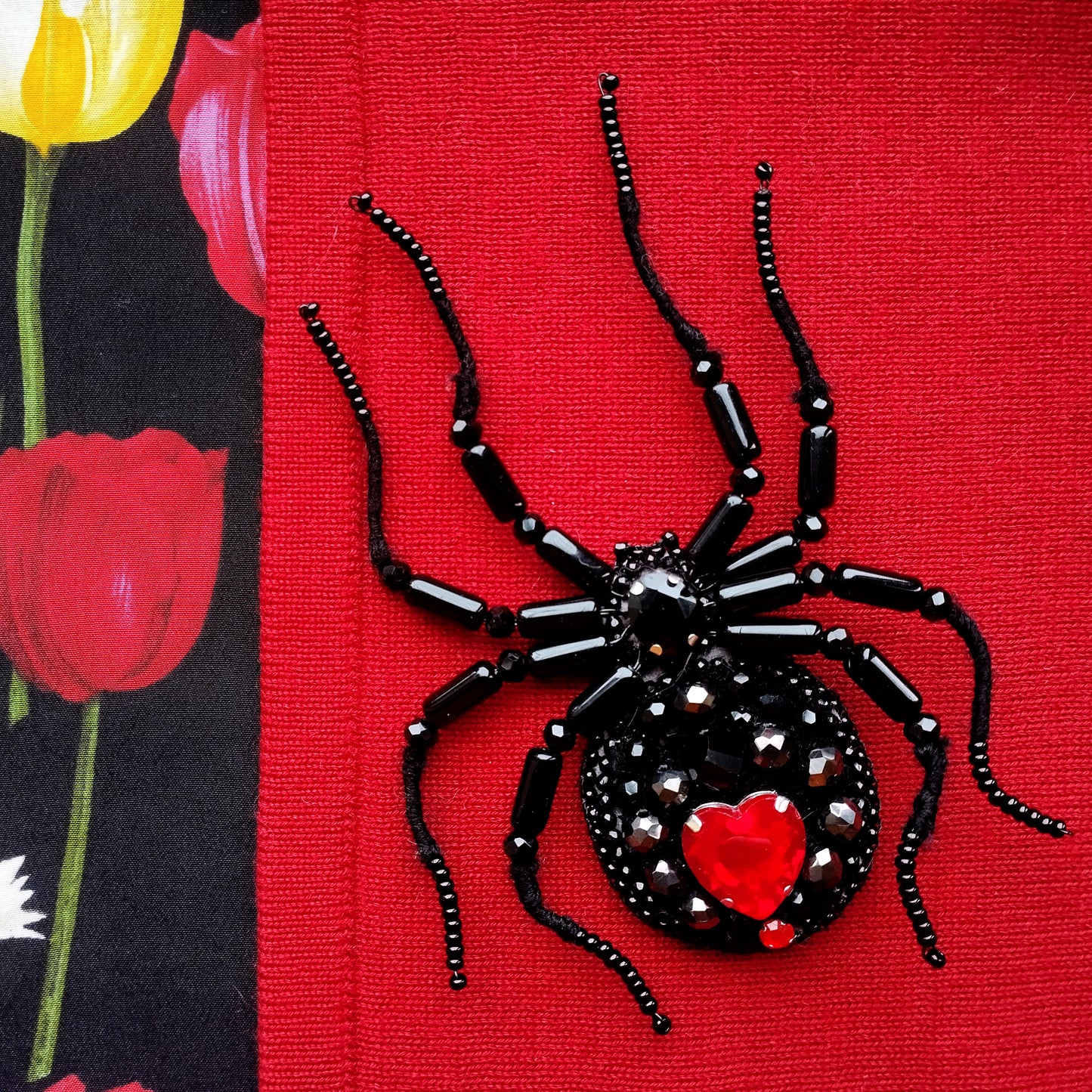 A close-up image of a black spider-shaped beaded piece with a red heart at its center, set against a red background.