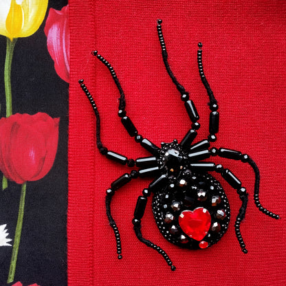A close-up image of a black spider-shaped beaded piece with a red heart at its center, set against a red background.