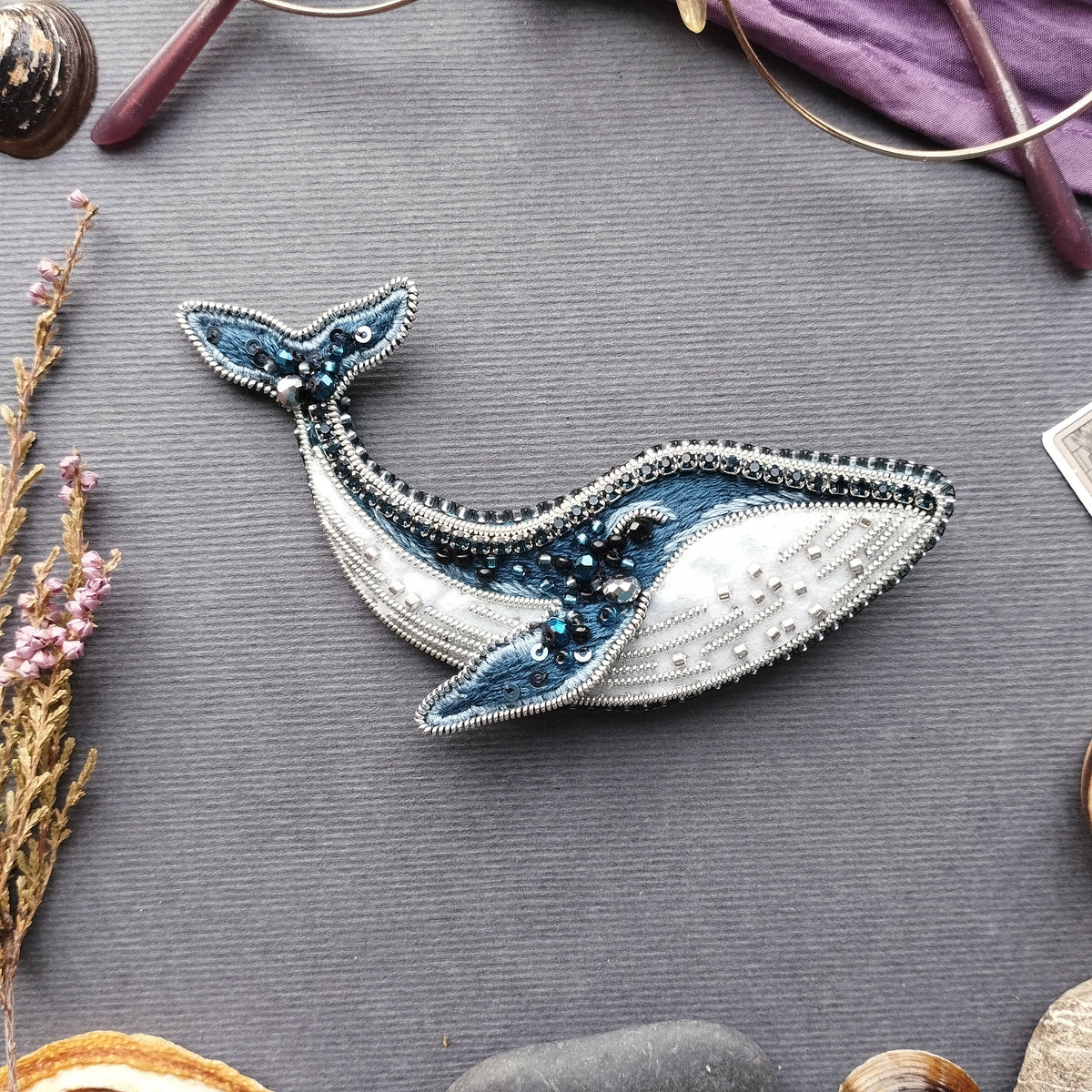 a handcrafted beaded and embroidered piece of art resembling a whale, placed on a gray surface surrounded by various objects such as a pair of glasses, a flower, and a rock.