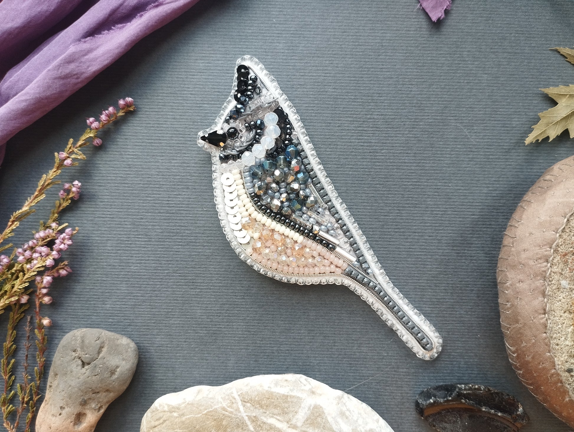 a close-up of a decorative bird embroidered piece, surrounded by various natural elements such as flowers, rocks, and leaves.