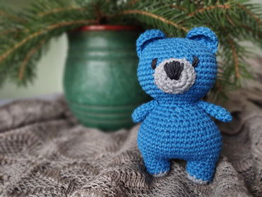a crocheted blue teddy bear sitting next to a pine tree