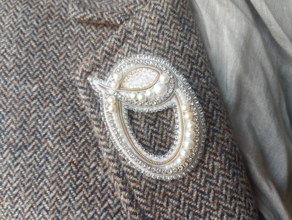 A close-up image of a decorative brooch or pin with pearls and a beaded design, attached to a tweed jacket or fabric.