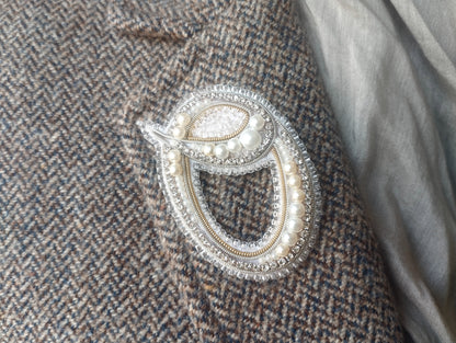 A close-up image of a decorative brooch or pin with pearls and a beaded design, attached to a tweed jacket or fabric.