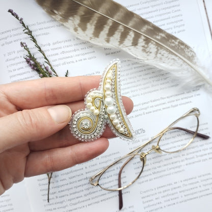 A hand holding a decorative brooch with pearls and a gemstone, surrounded by a feather, glasses, and a piece of paper with text.