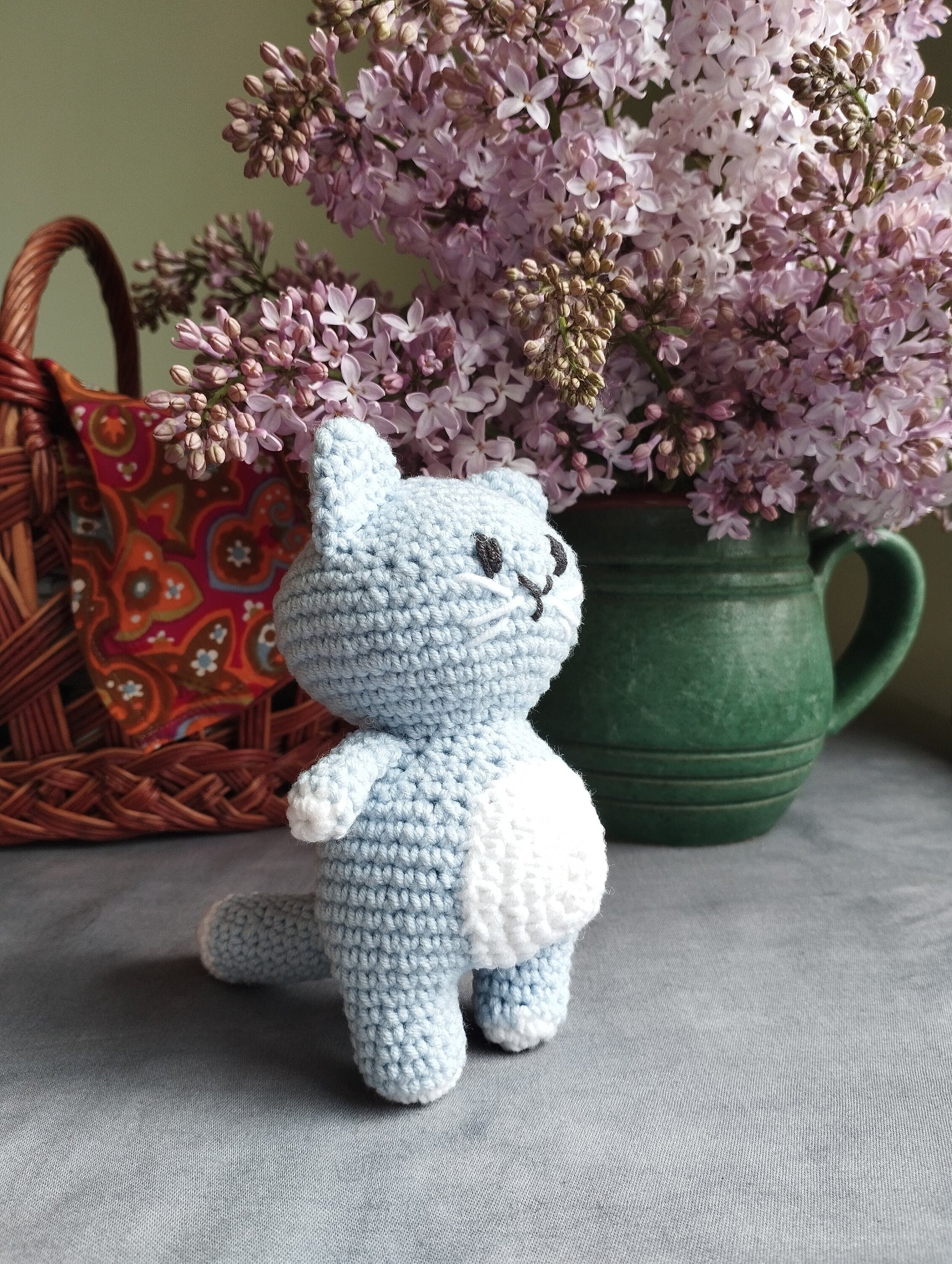 a crocheted cat sitting next to a basket of lilacs