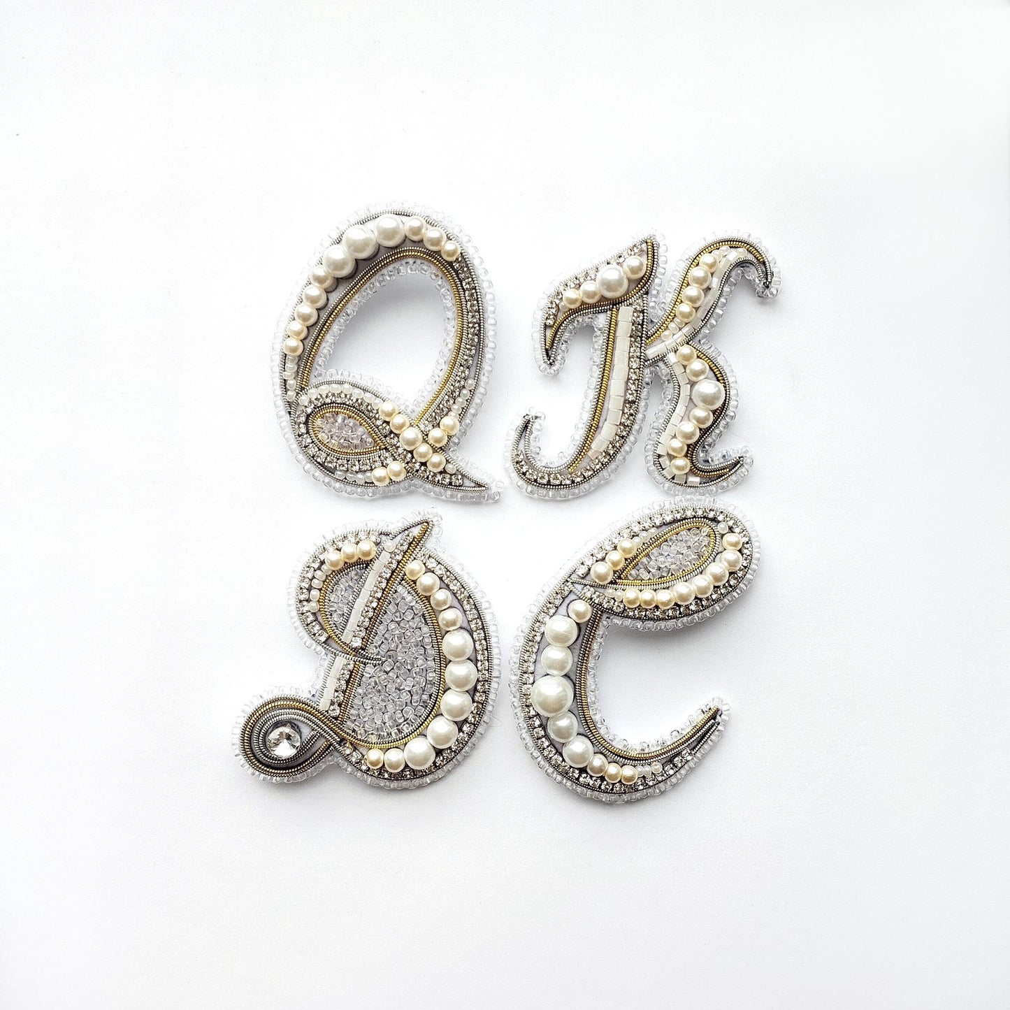 four ornate letters, each adorned with pearls and rhinestones, arranged in a square formation.