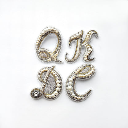 four ornate letters, each adorned with pearls and rhinestones, arranged in a square formation.