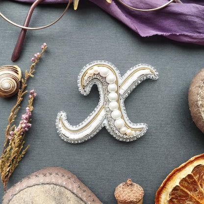a close-up view of a decorative letter 'X' made of beads and pearls, surrounded by various objects such as a pair of glasses, a seashell, a piece of fruit, and a small acorn.