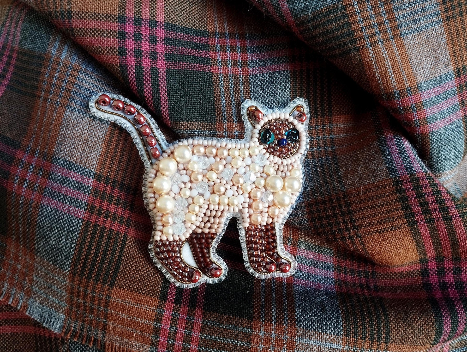 A decorative cat brooch made of beads and pearls is displayed on a plaid fabric background.