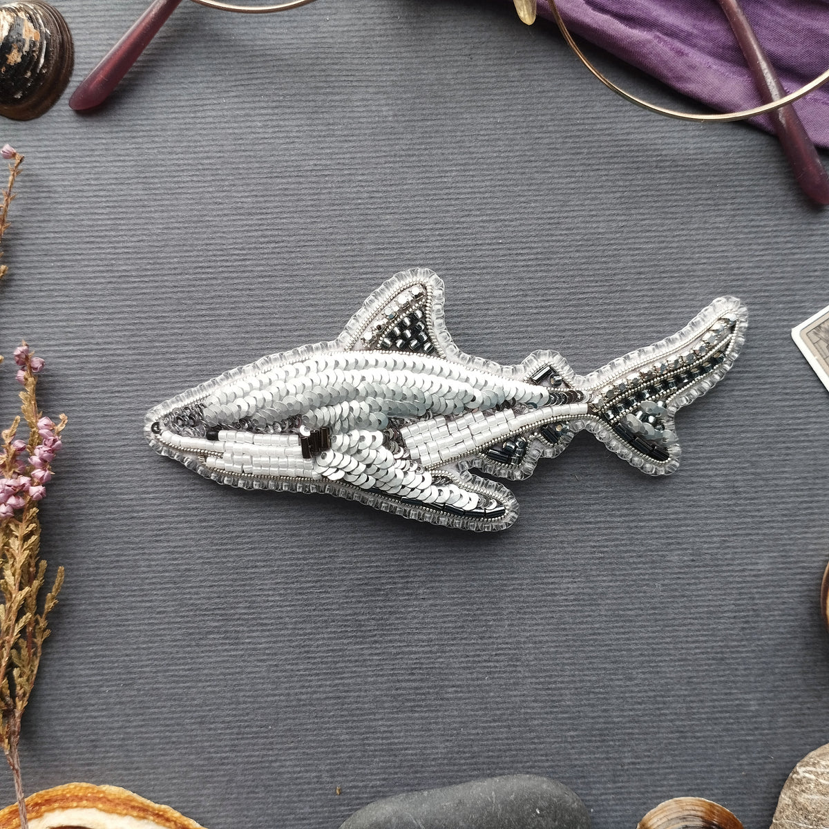 a detailed embroidered shark embroidery piece on a grey background, surrounded by various objects such as a pair of glasses, a flower, and a rock.