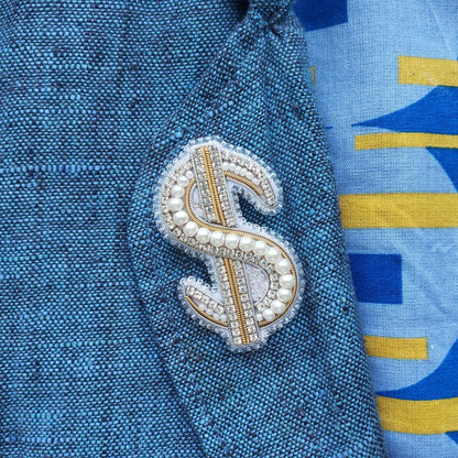 a close-up view of a blue fabric with a decorative dollar sign pin made of pearls and gold beads.