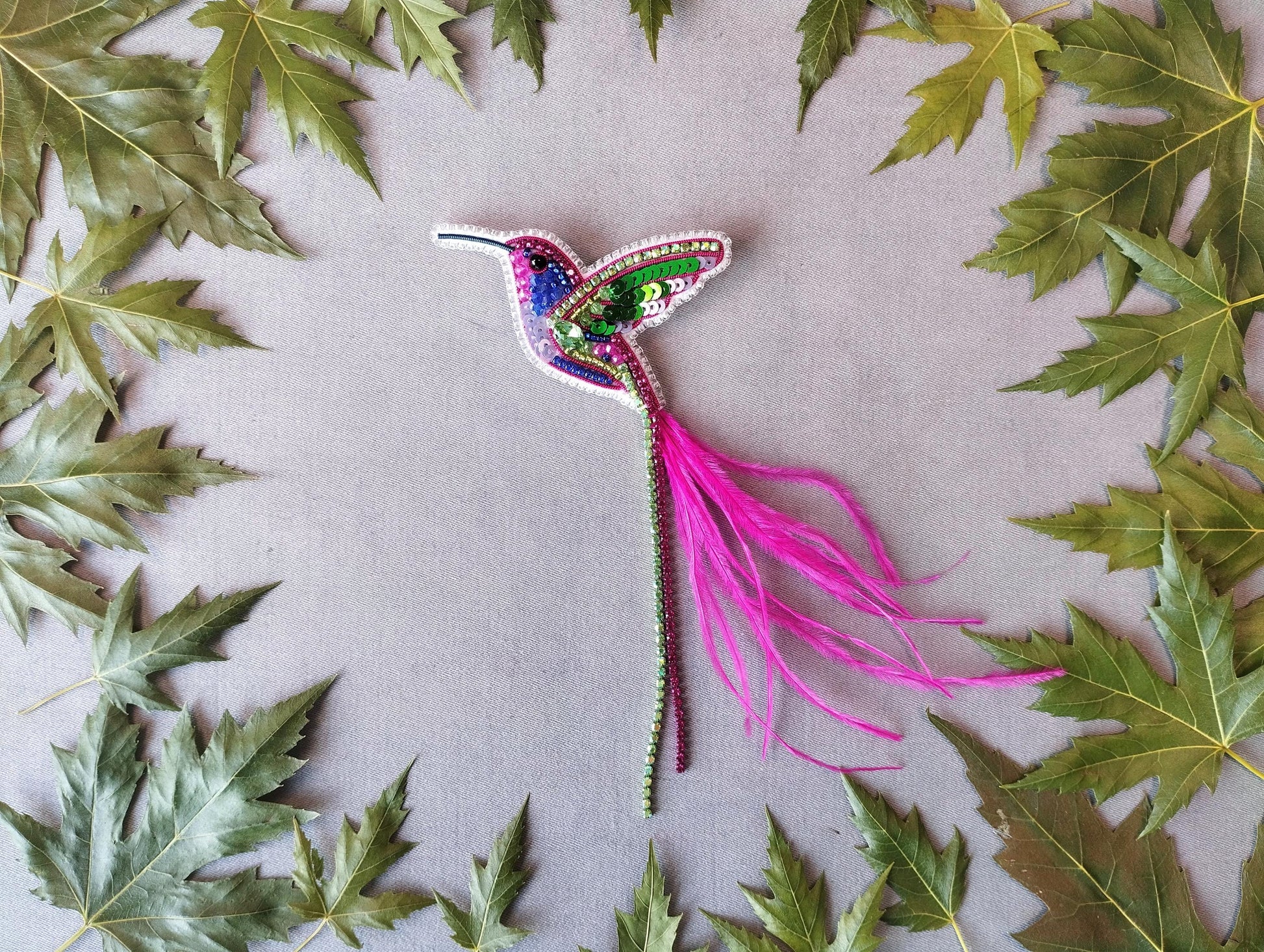 a colorful hummingbird brooch surrounded by green leaves on a gray background.