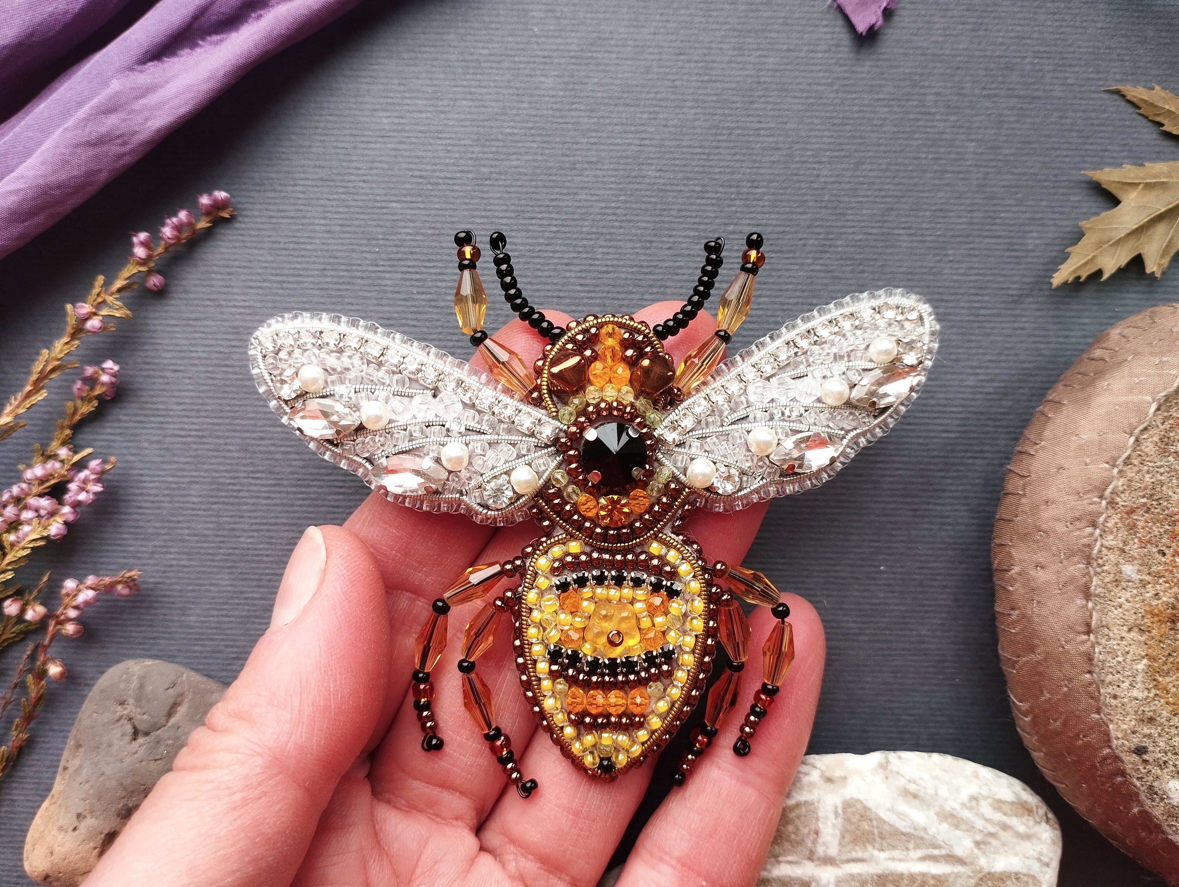 A hand is holding a decorative beaded insect brooch, which is intricately designed with various materials and colors.