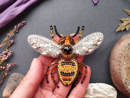 A hand is holding a decorative beaded insect brooch, which is intricately designed with various materials and colors.