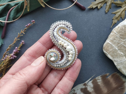 A hand holding a decorative letter 'S' made of silver, white, and blue beads, with a large crystal at the center.