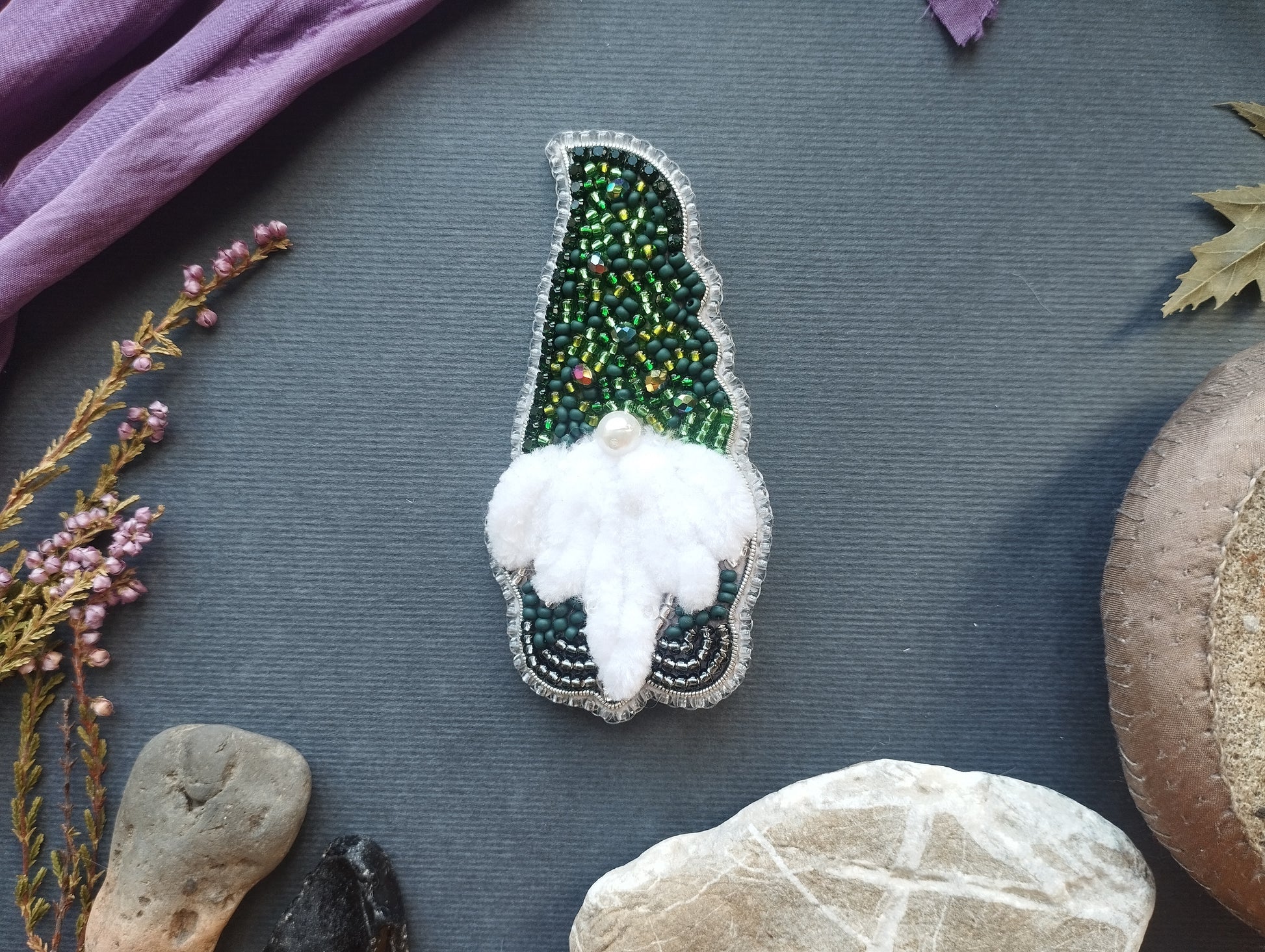 a close-up of a decorative item, possibly a brooch or pin, with a green and white color scheme and a white feather at the center. The item is surrounded by various natural elements, such as rocks, leaves, and flowers, creating a harmonious and visually appealing composition.