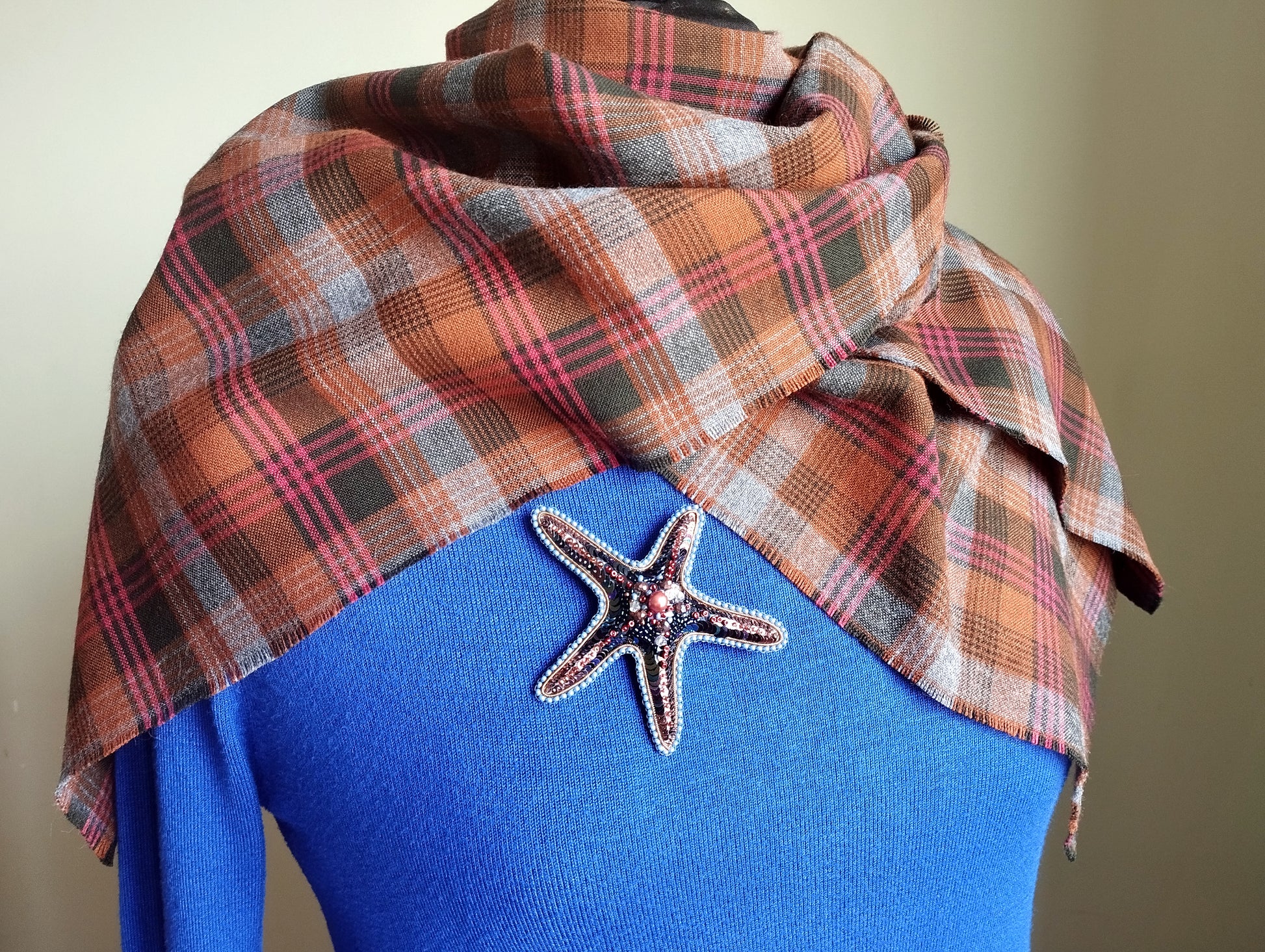 A plaid scarf is draped over a blue sweater, with a starfish brooch pinned to the sweater.
