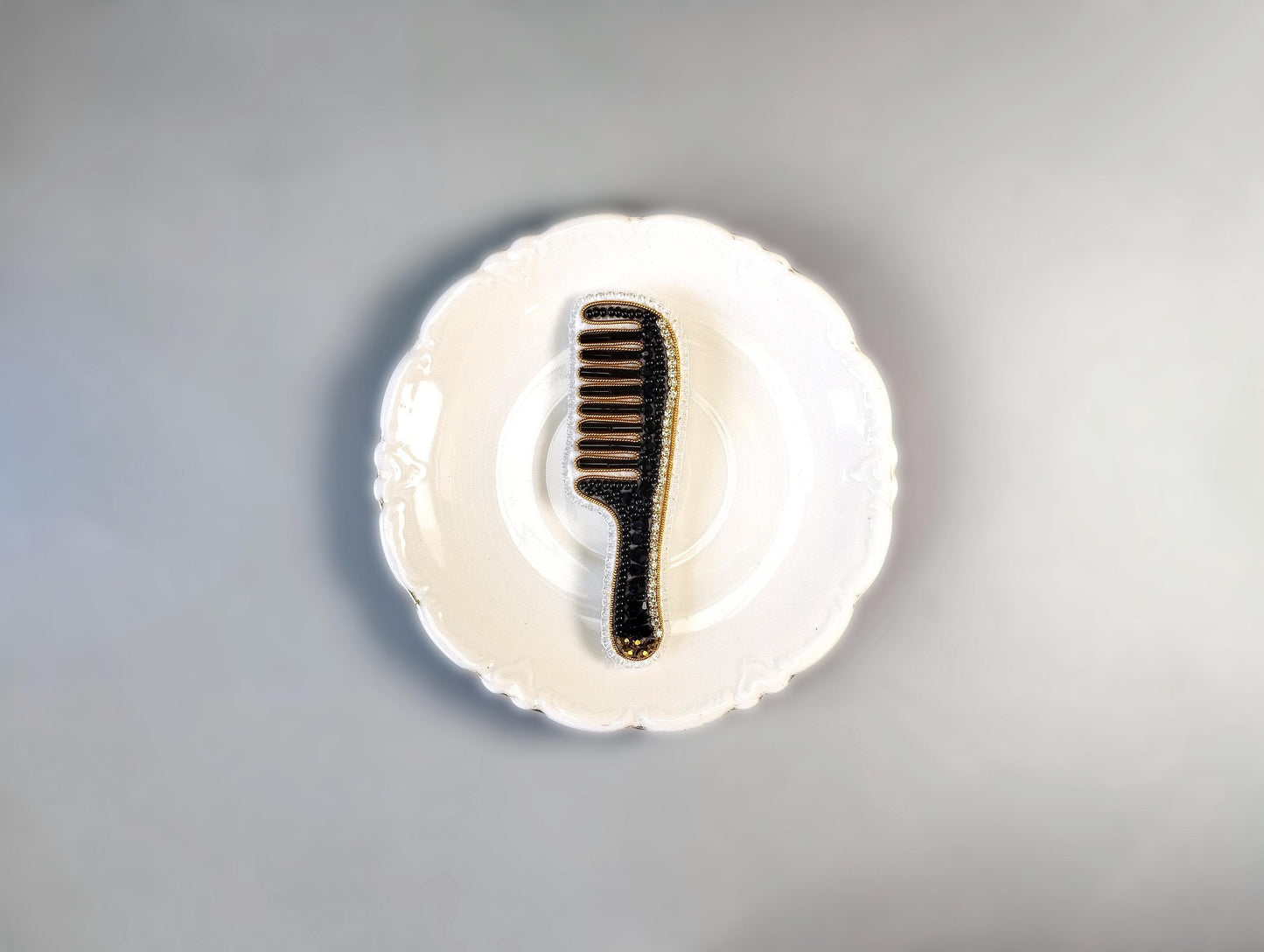 A black comb with gold accents is placed on a white, scalloped-edge plate against a gray background.