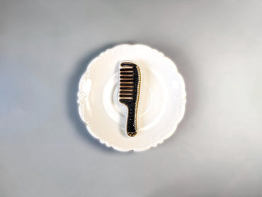 A black comb with gold accents is placed on a white, scalloped-edge plate against a gray background.
