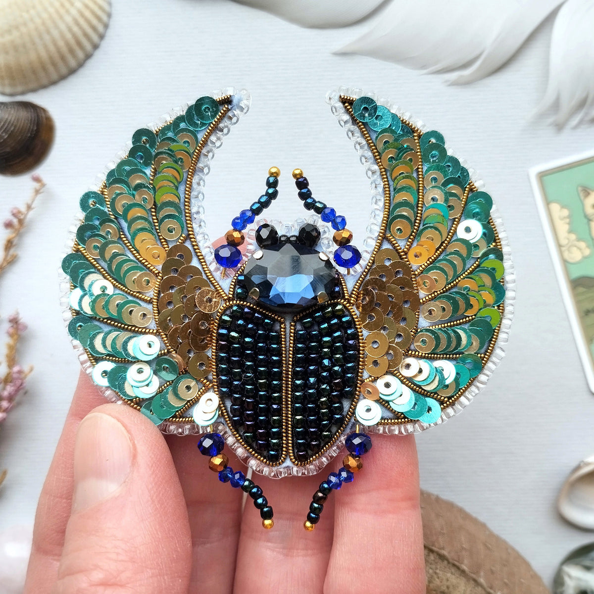 A hand is holding a decorative piece of jewelry featuring a detailed design of a large, colorful insect, possibly a beetle or a fly, with intricate patterns and sequins.