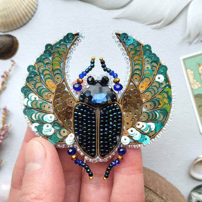 A hand is holding a decorative piece of jewelry featuring a detailed design of a large, colorful insect, possibly a beetle or a fly, with intricate patterns and sequins.