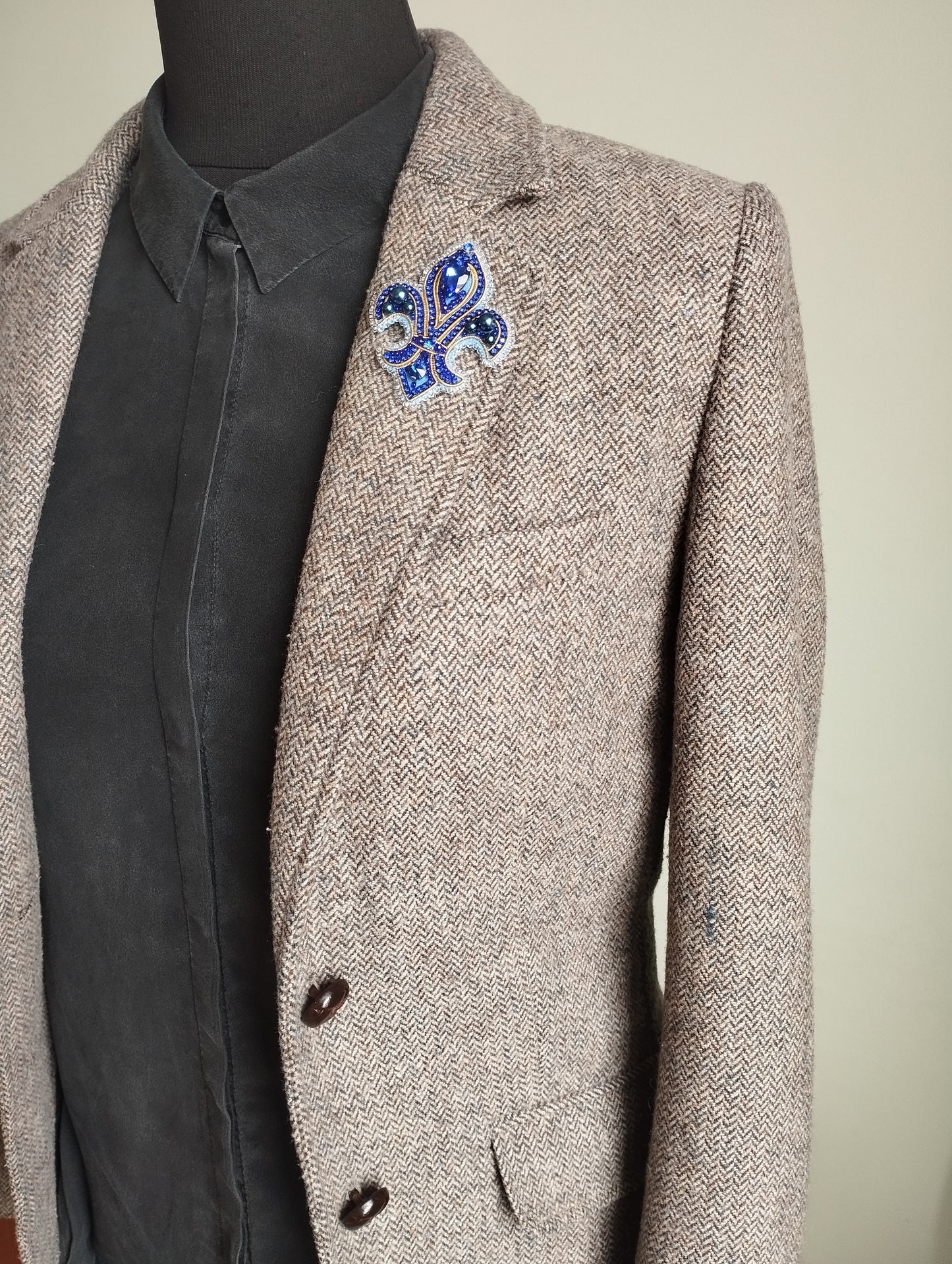 A mannequin wearing a gray tweed blazer with a blue and white brooch on the left side of the chest.