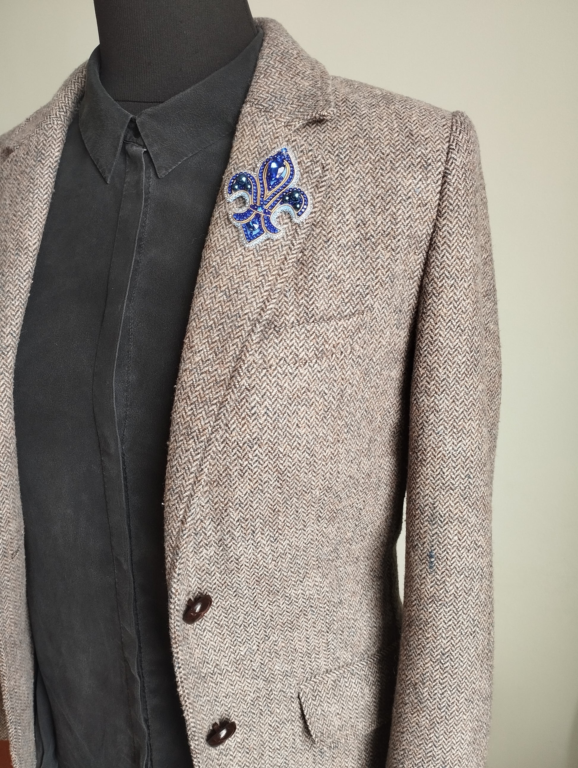 A mannequin wearing a gray tweed blazer with a blue and white brooch on the left side of the chest.
