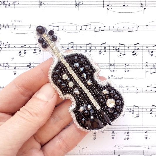A hand holding a decorative beaded object resembling a cello or violin, with sheet music visible in the background.