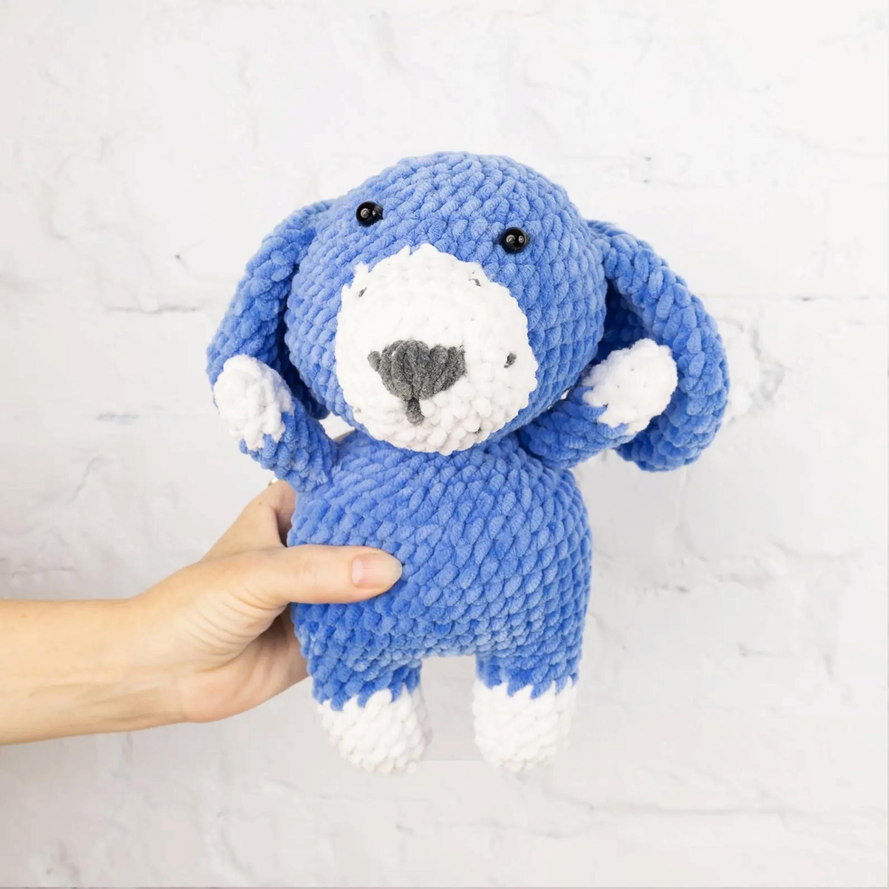 A hand is holding a blue and white crocheted stuffed animal, which appears to be a dog-shaped plush toy.