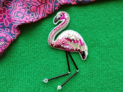A pink and white beaded flamingo brooch is displayed on a green surface, with a patterned fabric visible in the background.