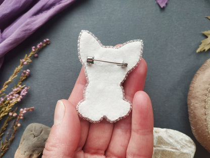 A hand holding a white felt cat-shaped pin against a dark background with various objects, including flowers and rocks.