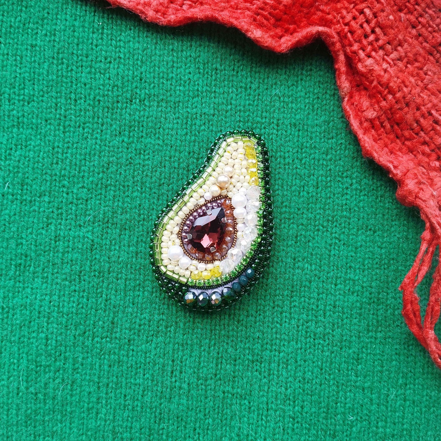 A beaded avocado-shaped brooch with a red gemstone is displayed against a green background, accompanied by a red fabric or cloth.