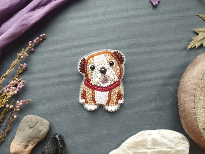 a small, intricately beaded dog brooch or pin placed on a grey background. Surrounding the dog are various objects, including a purple fabric, a leaf, and some rocks.