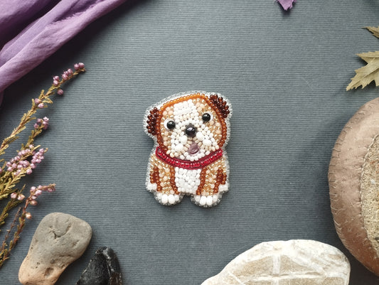 a small, intricately beaded dog brooch or pin placed on a grey background. Surrounding the dog are various objects, including a purple fabric, a leaf, and some rocks.