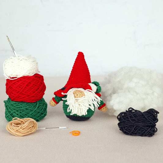 A crocheted gnome figurine with a red hat and green beard sits on a table surrounded by yarn, a crochet hook, and additional yarn balls.